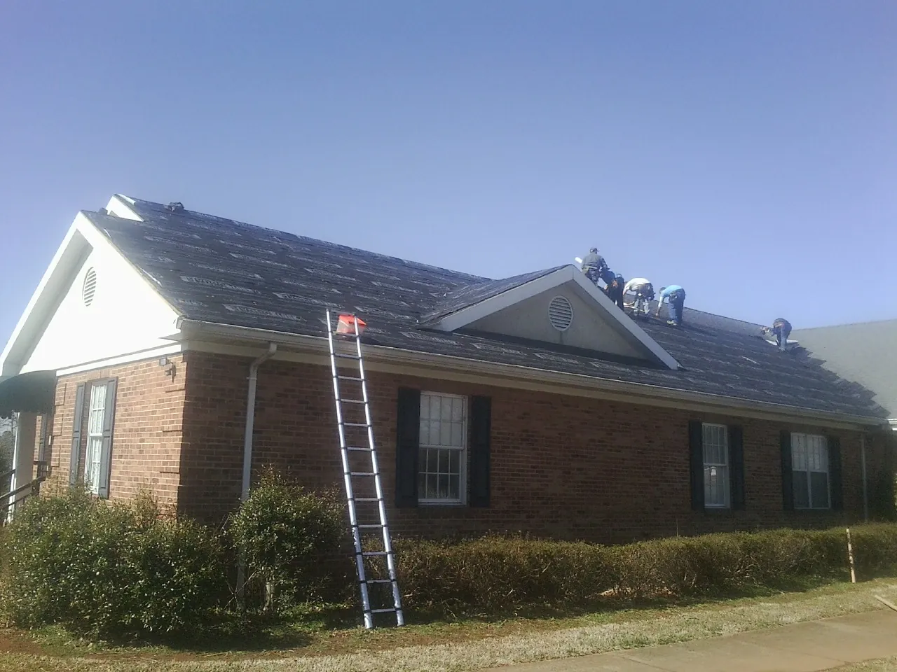 Crew tearing shingles off a brick ranch roof with a ladder set — Emergency Roof Repair work by our crew in Grafton